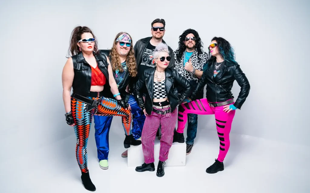 the Deloreans band posing in front of white background in neon and black leather costumes