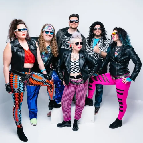 the Deloreans band posing in front of white background in neon and black leather costumes