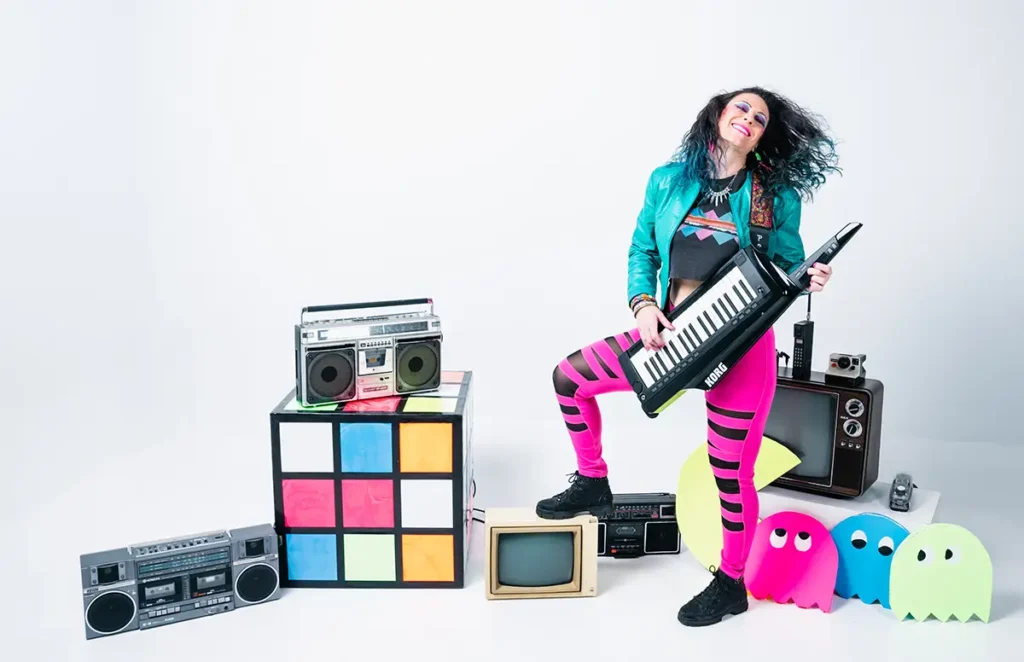 jennifer parker posing with foot on tv playing a keytar surrounded by 80s props
